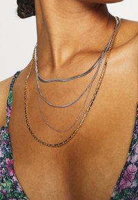 Gelaagde ketting met gouden en zilveren kettingen van verschillende texturen en ontwerpen, rustend op een sleutelbeen, met een top met bloemenpatroon.