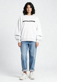 Felpa grigia con la scritta "MATCHA & DRAMA" in nero. Taglio ampio con spalle scese, abbinata a jeans azzurri a vestibilità rilassata e sneakers bianche.