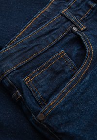 Tissu en denim bleu foncé avec des coutures orange, présentant une poche latérale et un détail de bouton métallique. Surface texturée avec un léger délavage.