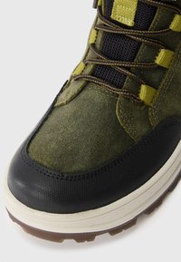 Botină de drumeție din suede verde, cu vârf din cauciuc negru, panou din plasă, șireturi oliv și talpă texturată. Include cusături și accente întărite.