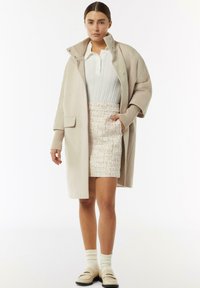 Beżowy wełniany płaszcz z wysokim kołnierzem i prążkowanymi mankietami, zestawiony z teksturowaną wiosenną spódnicą w pastelowych odcieniach. Modelka nosi kremowe skarpetki i płaskie beżowe buty.