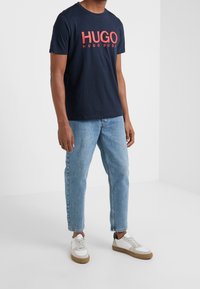 Námořnické bavlněné tričko s červeným logem "HUGO", doplněné světle modrými džínami a bílými teniskami s hnědými detaily.