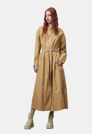 Abito camicia lungo in beige con vita con cintura, chiusura frontale a bottoni e due tasche laterali. Abbinato a stivali leggeri con lacci.