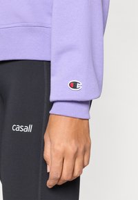 Μωβ φούτερ με μανσέτες ριμπ, στολισμένες με ταμπέλα λογότυπου; συνδυασμένο με μαύρο κολάν με λευκή επιγραφή. Ομαλή υφή υφάσματος.