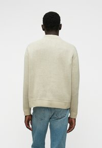 Pull en tricot beige avec un col rond, des poignets et un ourlet côtelés. Porté par-dessus un jean en denim bleu, avec un tissu texturé.