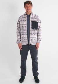 Giacca in fleece con zip e motivo tribale in grigio e bianco, con una tasca sul petto nera. Indossata con pantaloni grigi e Sneakers nere.