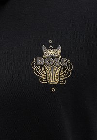 Melna auduma dizains ar zeltu izšūtu vārdu "BOSS" un smalkām detaļām ap to, ieskaitot kroni un rakstus.