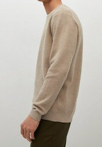 Pull en tricot beige à manches longues, avec un col rond et des poignets côtelés. Le tissu présente un motif texturé.