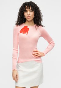 Ροζ μακρυμάνικη ριμπ μπλούζα με κόκκινο λουλουδάτο σχέδιο στον αριστερό ώμο και βολάν στα μανίκια· συνδυασμένη με λευκή υφασμάτινη φούστα.