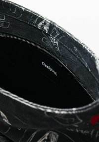 Mustast lõuendist koti sisemus, millel on valged visandid, hõbedane tõmblukk ja "Desigual" silt. Disain sisaldab erinevaid käsitsi joonistatud mustreid.