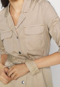 Beige skjortekjole med to brystlommer, knaplukning og bælte i taljen. Fremstillet af letvægtsstof med et struktureret design.