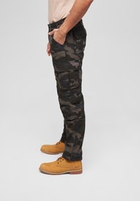 Camo cargo pants in dark green and black with multiple side pockets, worn with tan work boots, featuring a straight cut and durable material.