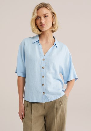 Žena s krátkymi svetlými vlasmi, oblečená v svetlomodrej košeli s krátkym rukávom a voľných béžových nohaviciach, stojaca s jednou rukou v vrecku.