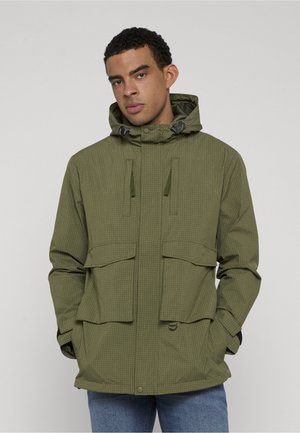 Młody mężczyzna ubrany w oliwkowozieloną kurtkę z kapturem i przednimi kieszeniami oraz niebieskie dżinsy, stojący na tle gładkiego szarego tła.