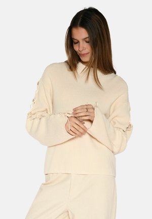 Cream ribbed sweater with a high collar, wide sleeves, and lace-up detailing along the sleeves. Smooth texture, relaxed fit.