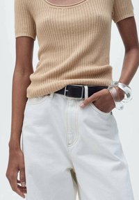 Mujer vistiendo una blusa de manga corta de canalé beige, pantalones blancos de talle alto con cinturón negro, y una gran pulsera transparente en la muñeca derecha, con la mano en el bolsillo.