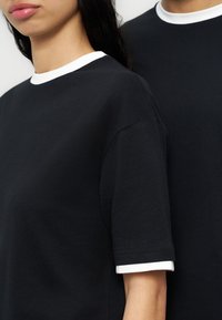 Δύο άτομα με μαύρα μπλουζάκια με λευκό λαιμόκοψη και μανσέτες στα μανίκια, εμφανίζονται από τους ώμους μέχρι τη μέση σε απλό φόντο.