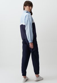 Giacca a vento azzurro chiaro e blu navy con polsini elasticizzati e una striscia bianca, abbinata a pantaloni jogger blu navy e scarpe da ginnastica bianche, mostrata di lato.
