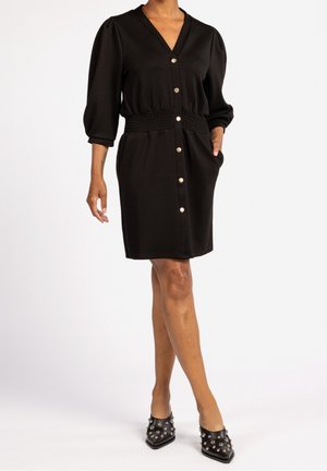 Kobieta w czarnej sukience zapinanej na guziki z trójczwartowymi bufkami na rękawach, marszczoną talią, bocznymi kieszeniami oraz czarnymi czółenkami z ćwiekami i ostrym noskiem.