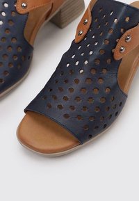Sandale en cuir perforé bleu marine avec des accents marron et un petit talon carré. Le design à bout ouvert présente des accessoires décoratifs et une semelle intérieure texturée.
