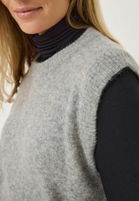 Gray sleeveless sweater made of soft, fuzzy fabric. Features a rounded neckline and raw edges at armholes, worn over a black turtleneck.
