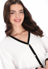 Witte cardigan met een zwarte V-hals en drie gouden knopen aan de voorkant. Zachte textuur, comfortabele pasvorm en lange mouwen.