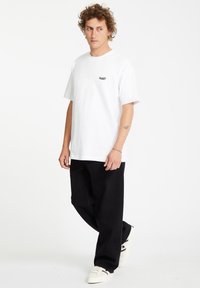 Weißes Baumwoll-T-Shirt mit kurzen Ärmeln, das ein kleines schwarzes Logo aufweist. Kombiniert mit lockeren, schwarzen Jeans und weißen Sneakern.