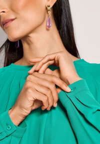 Grüne Bluse mit gefältetem Design und einem Knopfverschluss am Ärmelsaum. Abgerundet mit lila Tropfohrringen mit goldenen Akzenten.