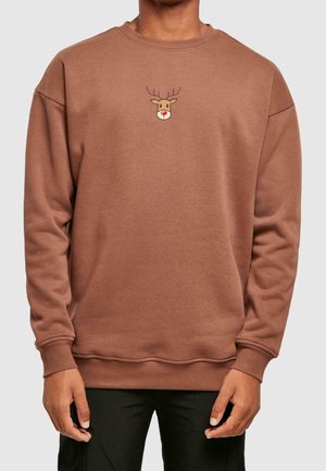Personne portant un sweat-shirt marron avec une petite illustration de visage de renne centrée sur la poitrine, associée à un pantalon noir.