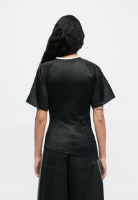 Hosszú fekete hajú személy, rövid ujjú fekete ingben és bő fekete nadrágban, háttal áll egy egyszínű háttér előtt.