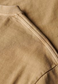 Beige cotton fabric with a smooth texture, featuring a rounded neckline and stitching along the edges for reinforcement.