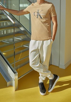 Osoba ubrana w beżowy t-shirt Calvin Klein Jeans, białe spodnie i czarne sneakersy stojąca przy metalowych i szklanych schodach wewnątrz.