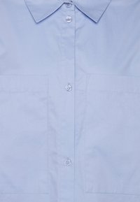 Camisa azul clara de botão, feita de tecido suave, com colarinho, dois bolsos frontais e fecho por botões ao longo do centro.