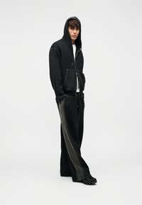 Μαύρο φούτερ με φερμουάρ και γκρι κουκούλα, σε συνδυασμό με μαύρο παντελόνι φαρδύ και χαρακτηριστικά υφασμένα μαύρα παπούτσια, τοποθετημένα σε απλό φόντο.