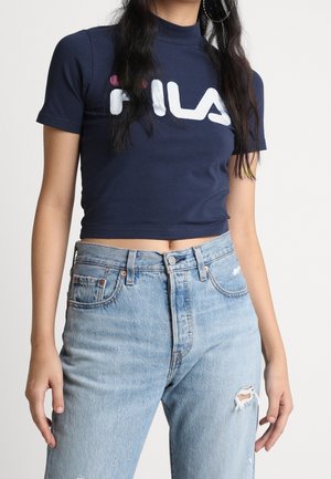 Vrouw draagt een marineblauw cropped FILA T-shirt en lichtblauwe high-waisted gebroken spijkerbroek, staand tegen een effen achtergrond.
