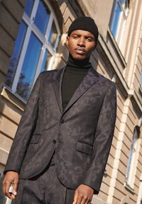 Chaqueta de traje de patrón oscuro en tonos negros, con dos botones frontales y pantalones a juego. Lleva una camiseta de cuello alto negra por debajo, exhibida al aire libre.