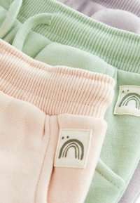 Tre par mjuka byxor i pastellfärger: rosa, grön och ljuslila. Varje par har en ribbad midja och en etikett med en regnbågsdesign.