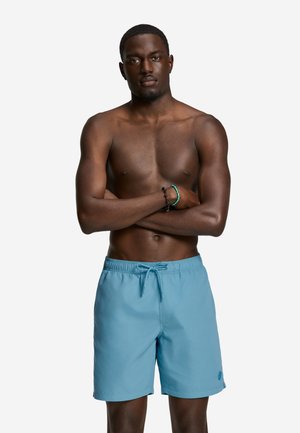 Bărbat musculos cu brațele încrucișate, purtând șorturi de baie bleu deschis și două brățări pe un fundal alb simplu.