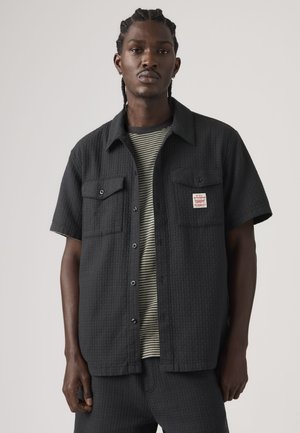 Joven con cabello trenzado que lleva una camisa negra de manga corta con botones sobre una camiseta de rayas blancas y negras.