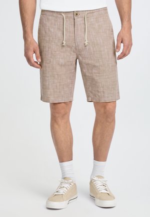 Modèle portant un short beige à carreaux avec cordon de serrage, une chemise blanche, des chaussettes blanches et des baskets beiges sur un fond uni.