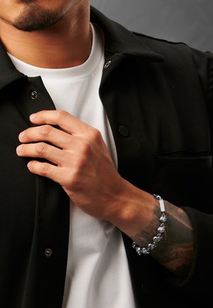 Silver bracelet with decorative links, worn on a wrist. Shirt is white, with a black, buttoned jacket partially visible. Textured skin.