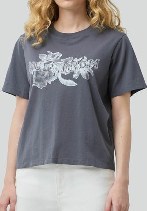 Kvinde iført en mørkegrå T-shirt med hvidt blomsterprint og teksten "MODSTRØM", kombineret med hvide bukser, foran en ensfarvet baggrund.