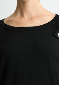 Haut en maille noire avec un col large côtelé, présentant des accents texturés aux épaules et un détail décoratif avec un bouton argenté sur le côté.