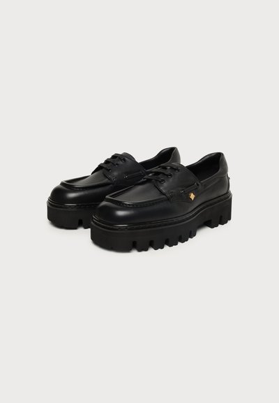 Paire de chaussures à plateforme noires épaisses à lacets avec semelles épaisses striées et petit accent doré sur le côté.