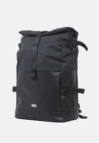 Zwarte rolltop rugzak gemaakt van waterdicht materiaal, met verstelbare schouderbanden, een strak ontwerp en een opvallend logodetail aan de voorkant.