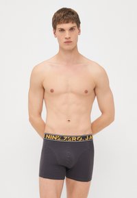 Schwarze Baumwoll-Boxershorts mit einem gelben Markenlogo-Bund, figurbetontem Design und glatter Textur. Das Model steht mit den Händen hinter dem Rücken.