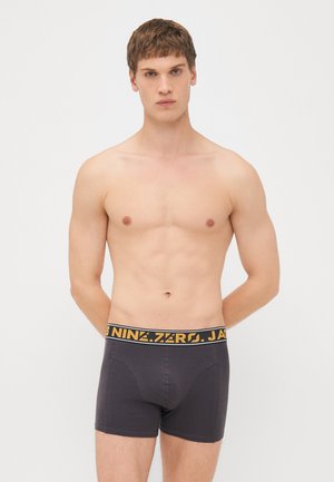 Black cotton boxer shorts with a yellow brand logo waistband, fitted design, and smooth texture. Model stands with hands behind back.