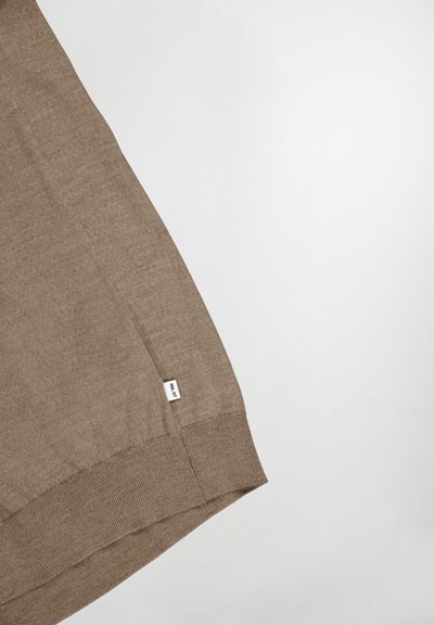 Bruine gebreide trui met een geribde zoom en een klein merklabel aan de onderkant. De stof lijkt zacht met een gladde textuur.