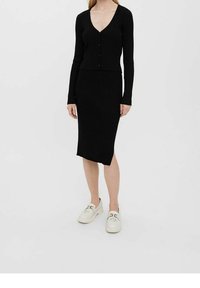 Mustahattu musta kahden osan asu, johon kuuluu pitkähihaista kardigania, jossa on napit, ja polven pituinen kapea hame, yhdistettynä valkoisiin kenkiin.