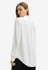 Camicia bianca a maniche lunghe realizzata in tessuto leggero, con colletto classico, orlo curvo e polsini con bottone. La parte posteriore presenta un dettaglio a yoke.